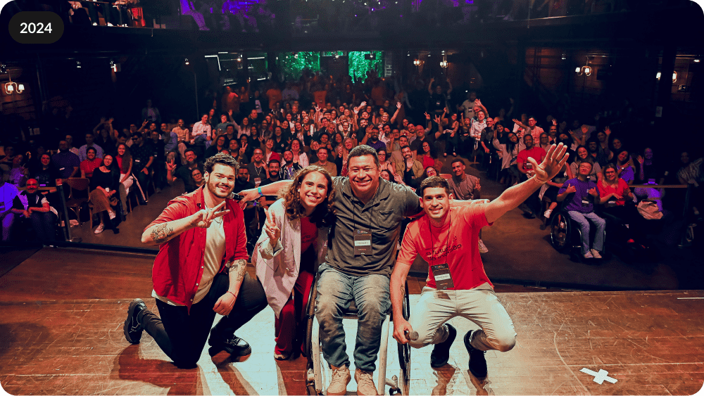 Foto do palco do Link 2024. Na imagem os mestres de cerimônia Roberto Castejon e Eduardo Victor estão com o atleta paralímpico Clodoaldo Silva e o CEO da Hand Talk Ronaldo Tenório. Ambos estão em cima do palco e de costas para a plateia. O público atrás está sentado e acena para a foto.