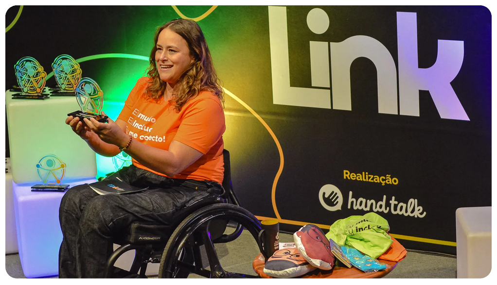 Foto do palco do Link 2023. Nela está a mestre de cerimônia Tabata Contri, uma mulher de pele clara e cabelos loiros. Sentada em sua cadeira de rodas, Tabata usa uma camiseta laranja do evento, uma calça jeans escura e segura um troféu sorrindo para a plateia. Ao seu lado esquerdo existem blocos com outros troféus e ao seu lado direito uma mesinha com brindes do evento. Ao fundo uma parede escura com o logo do Link. 