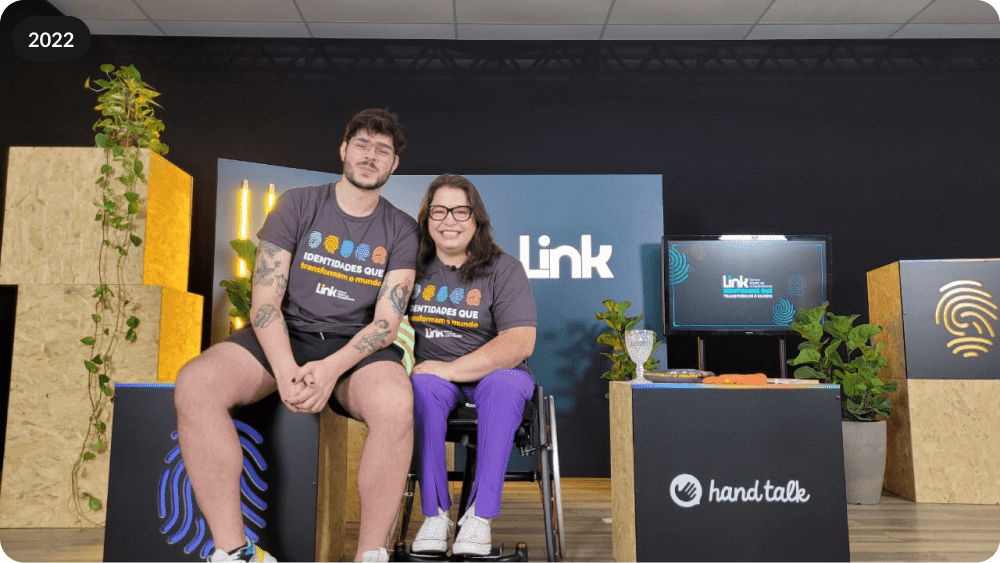  Foto dos mestres de cerimônia do Link 2022. Roberto Castejon e Carolina Ignarra estão no centro da imagem, envoltos ao cenário do evento com caixas que constam o logo do Link e da Hand Talk. Roberto, um homem branco de cabelos curtos castanhos, vestindo uma camiseta cinza e bermuda preta, está sentado em uma das caixas e sorri para a foto. Carolina uma mulher branca, de cabelos castanhos na altura do ombro, está sentada em sua cadeira de rodas vestindo ao lado esquerdo de Roberto, vestindo o mesmo modelo de camiseta cinza e uma calça roxa. 