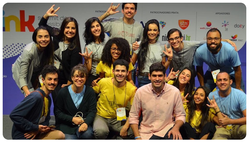 Foto da equipe Hand Talk no palco do Link 2019. Nela estão 15 pessoas sendo sete mulheres e oito homens. Todos sorriem para a foto. Ao fundo um painel com o logo do evento e de marcas patrocinadoras.