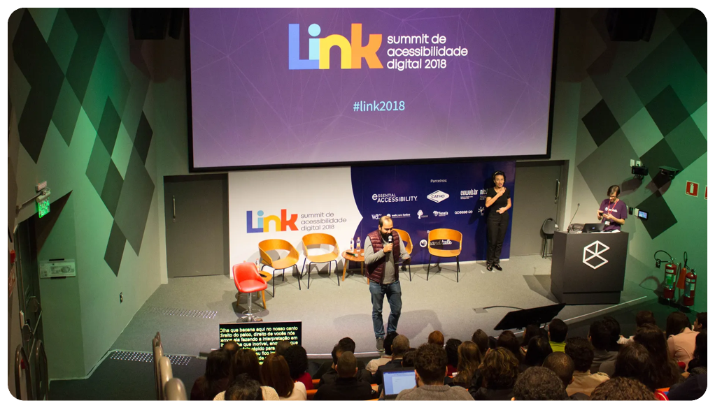  Foto do palco da edição de 2018 do Link. Ao centro da imagem existe um palestrante segurando o microfone e olhando para a plateia que está à frente dele. Ao lado esquerdo dele está a intérprete de Libras. Atrás dele existe um telão com o logo do evento ao centro. 