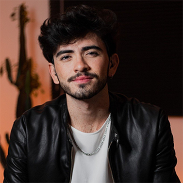 Gabriel é um homem branco de cabelos pretos, lisos e curtos. Ele usa barba. Gabriel olha para a câmera com um leve sorriso. Usa uma camiseta branca com jaqueta de couro preta. Ele usa uma corrente prateada.