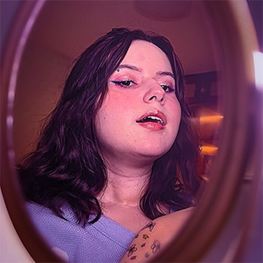 Marina é uma mulher de cabelos ondulados de cor escura. Ela se olha através de um espelho com a boca semiaberta. Ela usa uma maquiagem com olhos roseados, lápis de olho estilo “gatinho” e batom alaranjado.