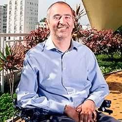 Fundo de um jardim com prédios atrás. Foto de homem branco com cabelos curtos, lisos e grisalhos. Ele está sorrindo, usando camisa azul e sentado em uma cadeira de rodas.