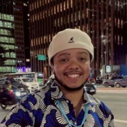 Akin Kolá, homem negro de boné bege e camisa estampada azul, sorrindo, com prédios e rua iluminada ao fundo.