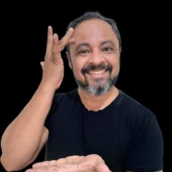 Alcelio Felix, homem de cabelos grisalhos curtos e barba, sorrindo e fazendo um sinal com as mãos, vestindo camiseta preta, sobre fundo preto