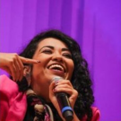 Amanda Lyra, mulher de cabelos cacheados escuros, sorrindo e segurando um microfone, vestindo blazer rosa, em palco com fundo roxo.