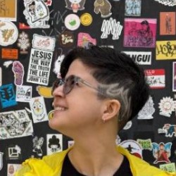 Ana Cuentro, pessoa de cabelos curtos e óculos, olhando para o lado, vestindo jaqueta amarela, em frente a uma parede de adesivos.