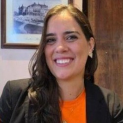 Ana Victorazzi, mulher branca de cabelos longos castanhos, sorrindo, vestindo blazer preto e camiseta laranja.