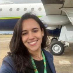 Andrea Paceli, mulher branca de cabelos longos escuros, sorrindo, com cordão verde no pescoço, em frente a um avião.