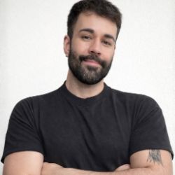 Bruno Antun, homem branco de cabelos castanhos curtos e barba, de braços cruzados, vestindo camiseta preta, sobre fundo claro