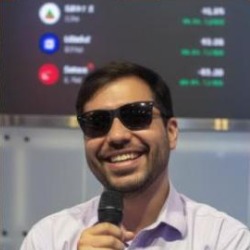 Bruno Simão, homem de cabelos curtos escuros e óculos escuros, sorrindo e segurando um microfone, vestindo camisa clara.