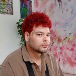 cabeça de chuva, homem de cabelos cacheados tingidos de vermelho e barba, vestindo camisa marrom, em frente a uma parede com pinturas.