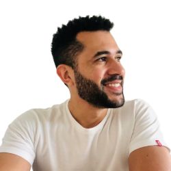 Davi Oliveira, homem de cabelos pretos curtos e barba, sorrindo e olhando para o lado, vestindo camiseta branca, sobre fundo branco