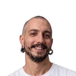 Felipe Carriço, homem branco careca e de barba, sorrindo, com alargadores nas orelhas, vestindo camiseta branca, em frente a um fundo branco.