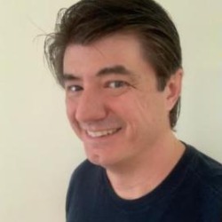 Guilhermo Reis, homem branco de cabelos castanhos, sorrindo, vestindo camiseta azul-marinho, em frente a uma parede clara.