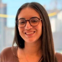 Jany Cavalcante, mulher branca de cabelos longos castanhos e óculos, sorrindo, vestindo blusa marrom, com janela e prédios ao fundo.