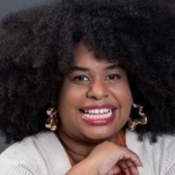 Juli Cintra, mulher negra de cabelos crespos volumosos, sorrindo, usando brincos dourados grandes.
