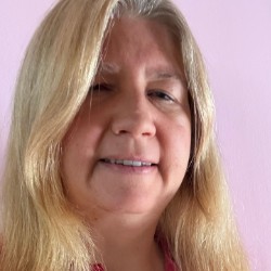 Karo Caran, mulher branca de cabelos loiros longos, sorrindo, vestindo camisa vermelha com bolinhas brancas, em frente a um fundo rosa-claro.