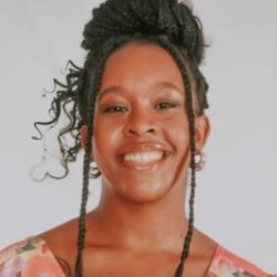 Lais Melo, mulher negra de cabelos cacheados presos e tranças, sorrindo, vestindo blusa estampada, em fundo cinza claro.