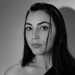 Leticia Fabbri, mulher de cabelos pretos longos, com expressão serena, vestindo blusa preta de um ombro só, em foto em preto e branco.