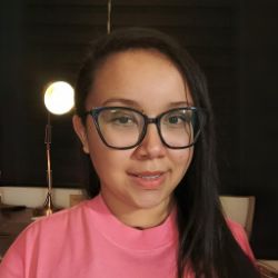 Liliane Claudia, mulher de cabelos pretos longos e óculos, sorrindo levemente, vestindo blusa rosa, em ambiente interno com uma luz ao fundo