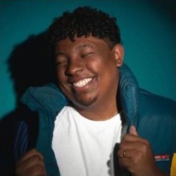 Marcos Vinni, homem negro de cabelos curtos cacheados, sorrindo de olhos fechados, vestindo jaqueta azul e camiseta branca.