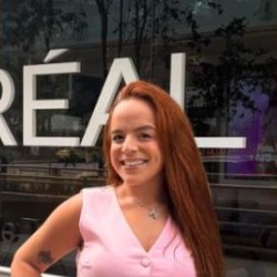 Nana Datto, mulher branca de cabelos longos ruivos, sorrindo, vestindo conjunto rosa, em frente à fachada da L’Oréal.