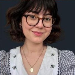 Natália Grein, mulher branca de cabelos castanhos com franja e óculos redondos, sorrindo, vestindo blusa branca bordada.
