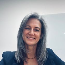 Paula Salomão, mulher branca de cabelos longos grisalhos, sorrindo, vestindo blazer azul-marinho, em ambiente interno