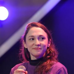Sara Paulo, mulher branca de cabelos ruivos ondulados, sorrindo e olhando para o lado, vestindo camiseta roxa e segurando um microfone, em um palco com iluminação roxa