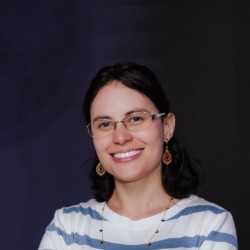 Sueidy Pires, mulher branca de cabelos pretos na altura dos ombros e óculos, sorrindo, vestindo blusa listrada branca e azul, em frente a um fundo escuro.
