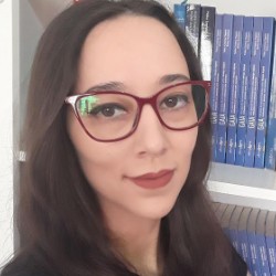 Talita Pagani, mulher branca de cabelos castanhos longos e óculos de armação vermelha, sorrindo levemente, em frente a uma estante com livros.