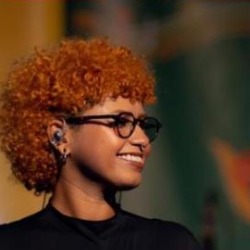 Thais Lobeu, mulher negra de cabelos cacheados ruivos e óculos, sorrindo de perfil, vestindo blusa preta.