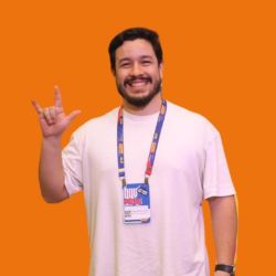 Thelmo Bulhoes, homem de cabelos pretos curtos e barba, sorrindo e fazendo um sinal com a mão, vestindo camiseta branca e crachá, sobre fundo laranja