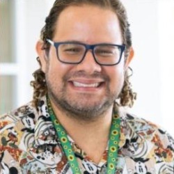 Thierry Cintra Marcondes, homem branco de cabelos cacheados, óculos e barba, sorrindo, vestindo camisa estampada e cordão verde.